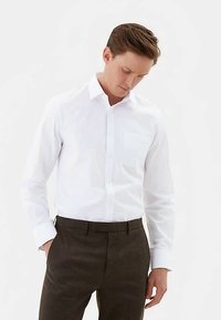 Hvid skjorte med knapper, krave og enkelt brystlomme, kombineret med teksturerede, mørkebrune ternede bukser. Struktureret pasform og glat stof.