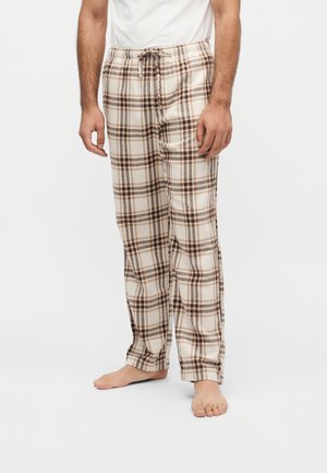 Homme portant un pantalon de pyjama à carreaux beige et marron avec une taille à cordon, debout pieds nus sur un fond blanc uni.