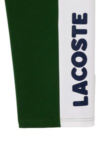 Zöld pamut rövidnadrág, fehér oldalsó panellel, amelyen a "LACOSTE" logó sötétkék színben látható. Sima tapintású, laza fazon.