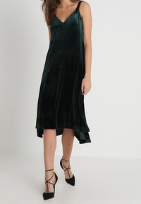 Grünes Samtkleid mit V-Ausschnitt und verstellbaren Trägern, mit asymmetrischem Saum und glatter Textur. Kombiniert mit schwarzen Riemchen-High Heels.