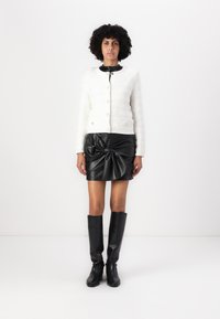 Veste blanche texturée avec un col rond, associée à une jupe en cuir noir avec un design noué, et des bottes noires à hauteur de genou.