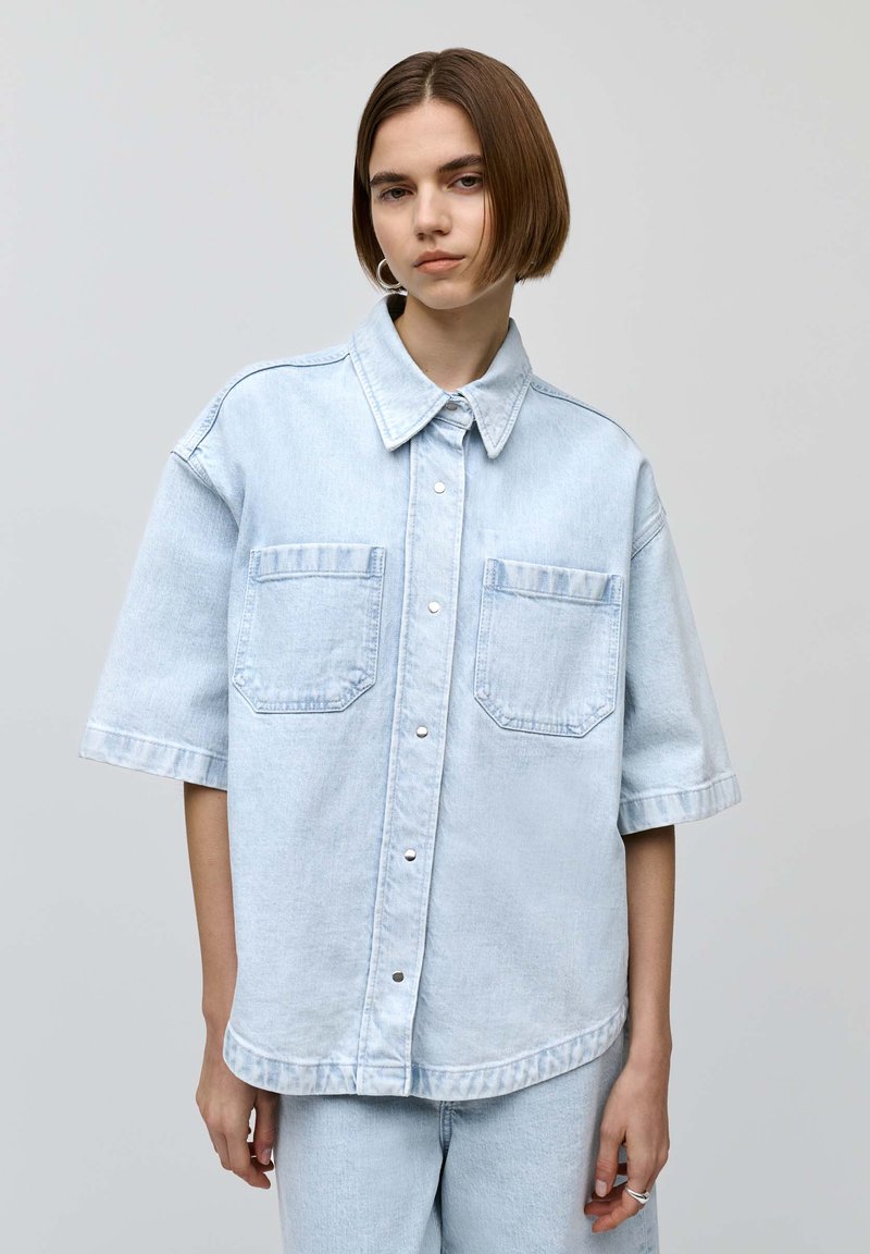 Camisa de mezclilla de un suave color azul claro con botón frontal, cuello puntiagudo, mangas cortas y dos bolsillos en el pecho. Con un tejido suave y un diseño de corte relajado.