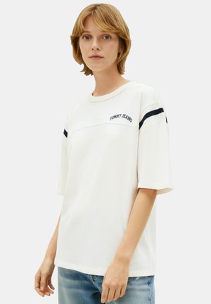 Giovane che indossa una maglietta bianca Tommy Jeans con righe nere sulle maniche e jeans azzurri, in piedi davanti a uno sfondo neutro.