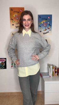 Žena se usmívá, má ruce v bok, na sobě šedý svetr s copánkovým vzorem přes žlutou košili na knoflíky, stojí v místnosti s barevnými plakáty a knihami.