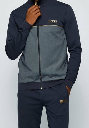 Homme portant un survêtement Hugo Boss bleu marine et gris, fermeture éclair partiellement fermée, une main près du col et l'autre dans la poche.