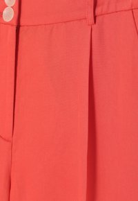 Primer plano de una tela color coral, con un patrón texturizado tejido y dos botones grandes en la cintura. Se aprecia un pliegue limpio y bien ajustado.