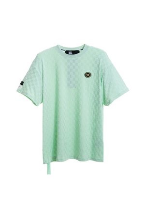 Persona rubia con camiseta verde claro texturizada de gran tamaño y pantalones cortos a juego con ribete negro, calcetines blancos y zapatos beige texturizados sin cordones.