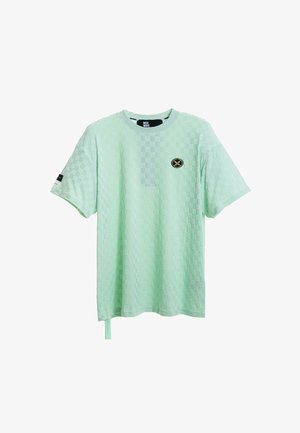 Persona rubia con camiseta verde claro texturizada de gran tamaño y pantalones cortos a juego con ribete negro, calcetines blancos y zapatos beige texturizados sin cordones.