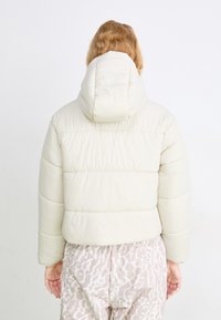 Giacca beige gonfiabile con cappuccio, featuring sezioni orizzontali cucite e una vestibilità rilassata, abbinata a pantaloni leggeri a fantasia.
