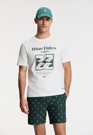 Camiseta blanca de algodón con estampado de "BlueTides Resort", combinada con pantalones cortos verdes con un sutil patrón de hojas, y una gorra verde con un logo.