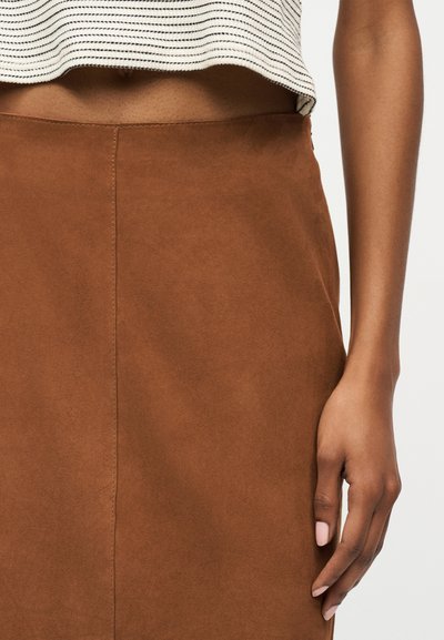 Κλείσιμο πλάνου ενός ατόμου που φοράει καφέ σουέτ φούστα και κοντό λευκό-μαύρο ριγέ τοπ, με ένα χέρι χαλαρωμένο δίπλα στο σώμα.