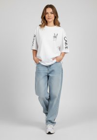 Balta per didelė t-shirt su rankų grafika ir užrašais "HAVE" ir "CARE" ant rankovių, derinama su šviesiai mėlynomis plačiomis džinsomis ir baltomis sportinėmis bateliais.