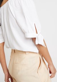 Camicia bianca con maniche a sbuffo, dettagli plissettati e rifiniture a fiocco sui polsini, abbinata a pantaloni beige chiaro con tasche posteriori.