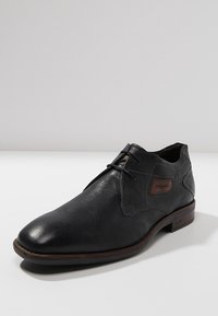 Chaussure habillée en cuir noir avec une finition lisse, bout rond et coutures discrètes. Présente un accent en cuir marron sur le côté et un talon bas.