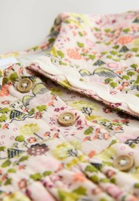 Kvetinová látka v pastelovej ružovej farbe, vybavená gombíkmi a vlnitou lemovkou. Dizajn zahŕňa rôzne tvary a farby, predovšetkým zelené a oranžové odtiene.