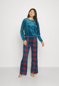 Haut en velours teal à manches longues associé à un pantalon à jambes larges à motifs plaid avec des couleurs bleue, rouge et verte. Les deux vêtements ont une texture douce.