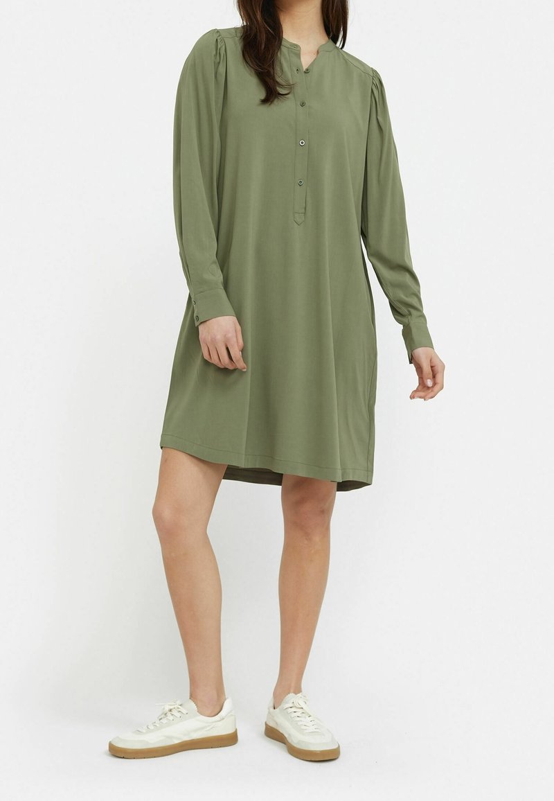 Robe olive, à manches longues, avec un col henley à boutons et des épaules froncées, fabriquée en tissu doux, associée à des chaussures blanches.