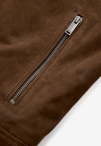 Tejido de ante marrón con un zipper lateral que presenta un tirador metálico plateado y dientes finos, destacando una textura suave y lisa.