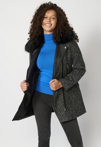 Crno-bijeli kaput s teksturom i crnim umjetnim krznom na ovratniku, s patent zatvaračem i bočnim zatvorenim džepovima, nosi se preko plave turtleneck majice.