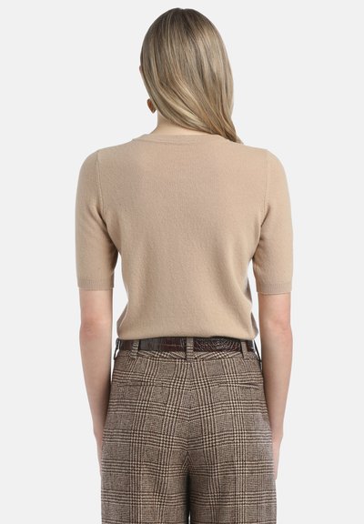 Bežs īsām piedurknēm svīteris ar apaļu kakla izgriezumu un mīkstu tekstūru, apvienots ar brūnām ģeometriskām augstām biksēm un plānu jostu.