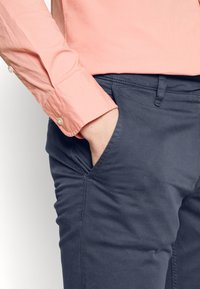 Hellrosa Langarmshirt mit Perlenknöpfen, kombiniert mit marineblauen Hosen mit Seitentasche und glatter Textur.