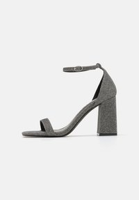 Grå teksturert høyhælt sandal med en bred stropp over tærne, ankelstropp med justerbar spenne, og et kraftig hældesign.