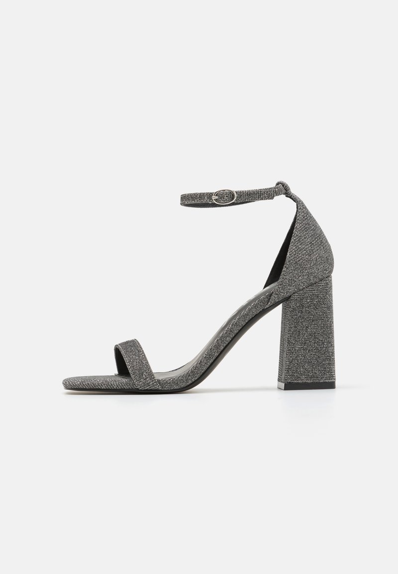 Grå teksturert høyhælt sandal med en bred stropp over tærne, ankelstropp med justerbar spenne, og et kraftig hældesign.