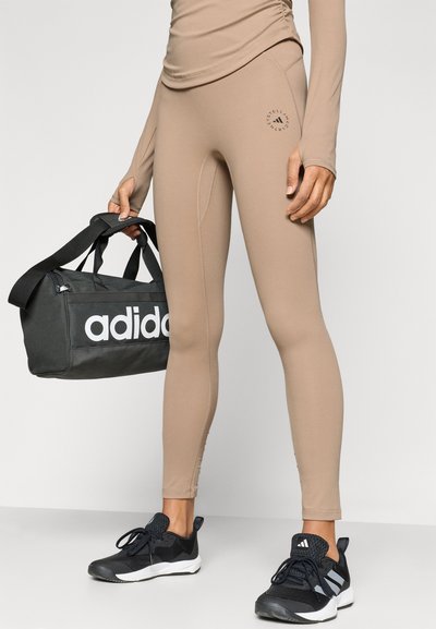 Μπεζ κολάν με εφαρμοστή σχεδίαση, με στάμπα λογότυπου στον μηρό; σε συνδυασμό με μαύρη τσάντα duffel και μαύρα αθλητικά παπούτσια.
