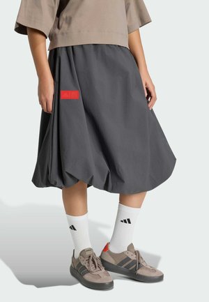 Persona vistiendo una falda globo gris con un parche rojo, zapatillas deportivas beige, calcetines blancos de Adidas y un top corto beige, mostrado desde la cintura hacia abajo.