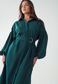 Tiffany-vihreä mekko, joka on valmistettu pehmeästä kankaasta. Mekossa on napitettu etuosa, vyötäröllä vyö ja puffatut pitkät hihat, joissa on sileä tekstuuri.