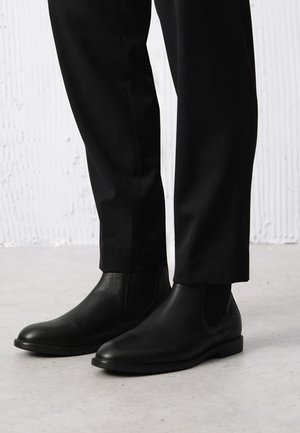 Czarne skórzane botki Chelsea z elastycznymi panelami bocznymi, gładką teksturą, zaokrąglonym noskiem i płaską podeszwą, w zestawieniu z czarnymi spodniami.