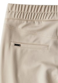 Beige Hosen mit elastischem Bund, einer abgesteppten Gesäßtasche und einem kleinen rechteckigen schwarzen Markenlabel am Taschenrand.