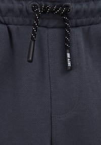 Tmavě šedé tepláky z texturované látky, s elastickým pasem a nastavovacími černobílými šňůrkami, a s malým logem.