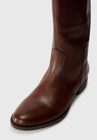 Brown leather knee-high boot with a pointed toe and stitched detailing, resting on a light background.
