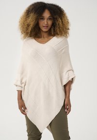Beige gestrickter Poncho mit diagonaler Rippstruktur, weit geschnittenen Ärmeln und V-Ausschnitt, kombiniert mit olivgrünen Hosen. Glatte Textur.