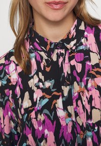 Blusa floreale nera, con un colorato motivo astratto nei toni del rosa, blu e beige. Collo a camicia con bottoni e dettagli con bottoni frontali.