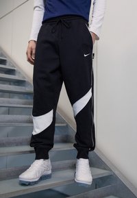 Pantalones de chándal negros con paneles de acento blancos, cinturilla elástica y bolsillos laterales, combinados con zapatillas blancas en una escalera.