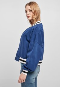 Chaqueta bomber azul de corte holgado, puños acanalados en negro y blanco, y cierre de botones a presión. Tela suave con textura lisa.