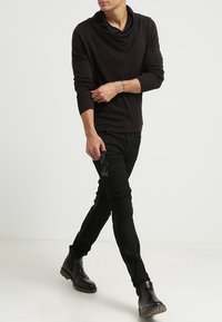 Svart långärmad topp med en krage, ihop med slim-fit svarta byxor och svarta kraftiga stövlar. Modellen håller i solglasögon.