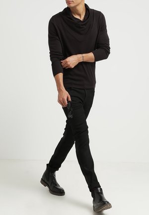 Schwarzes Langarmoberteil mit Kragen, kombiniert mit enganliegenden schwarzen Hosen und schwarzen klobigen Stiefeln. Model hält Sonnenbrille.