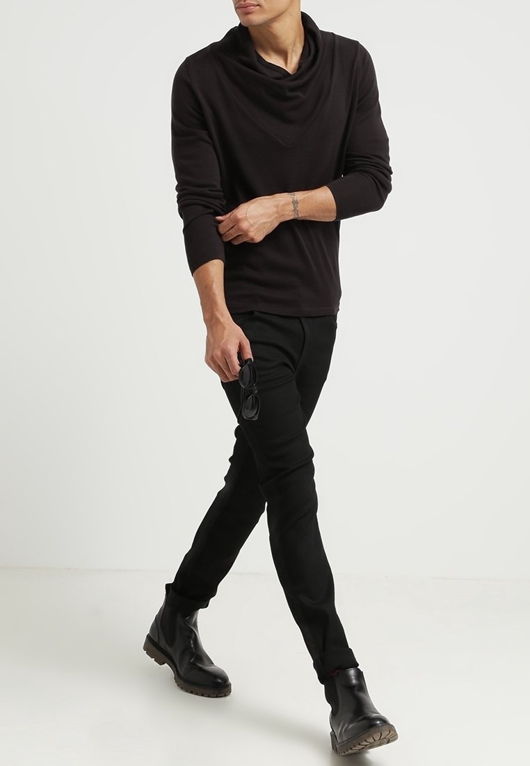 Svart långärmad topp med en krage, ihop med slim-fit svarta byxor och svarta kraftiga stövlar. Modellen håller i solglasögon.