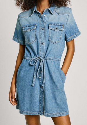 Vrouw die een korte mouw denim jurk met knopen aan de voorkant, borstzakken en een gestrikte tailleband draagt, staand met één hand in de zak.