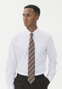 Balti apsiaustas marškiniai su ilgomis rankovėmis, derinami su ruduo kaklaraiščiu, kuris turi įstrižas violetines juostas. Tamsios kelnės užbaigia aprangą.