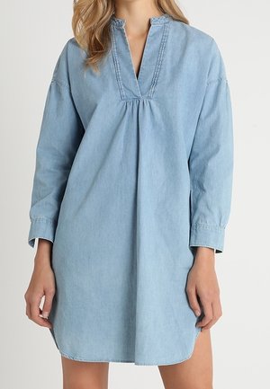 Kvinde iført en lysblå, langærmet denimkjole med V-udskæring og let opsamlet detalje foran.