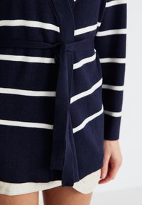 Cardigan azul-marinho com riscas brancas, feito de material de malha suave. Apresenta um cinto de laço na cintura e mangas longas.