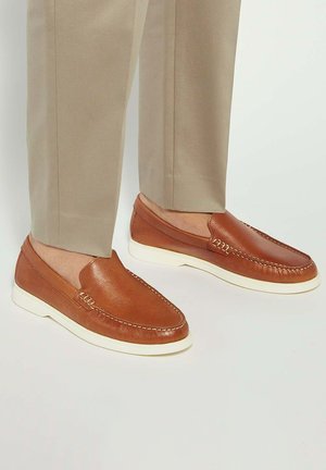 Brown leather loafers with white rubber soles, featuring stitching details and a smooth, shiny texture. Worn with light tan trousers.