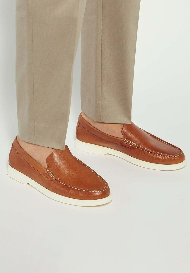 Brown leather loafers with white rubber soles, featuring stitching details and a smooth, shiny texture. Worn with light tan trousers.