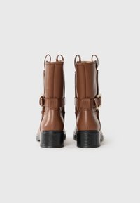 Botas de tobillo de cuero marrón con una textura suave, caña corta, tiradores traseros y un acento de hebilla lateral. Tacón negro apilado.