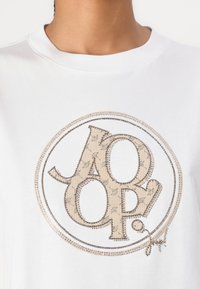 Weißes Baumwoll-T-Shirt mit einem runden Logo in Beige und Silber, das auf der Vorderseite mit kleinen Sternen gemustert ist.