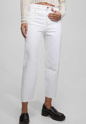 Henkilö, joka pukeutuu korkeavyötäröisiin valkoisiin lyhytlahkeisiin farkkuihin, joissa on repsottavat reunat, mustiin korollisiin loafereihin ja raidalliseen pitkähihaiseen lyhytpaitaiseen toppiin.